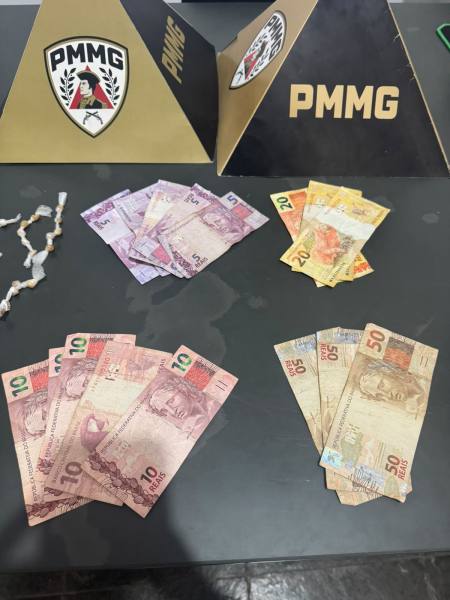 Notas de dinheiro em diferentes denominações dispostas em uma superfície, com cartazes da PMMG ao fundo.