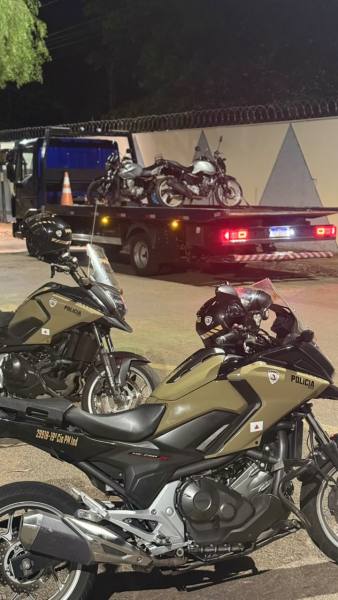 Duas motos de polícia estacionadas em primeiro plano, com uma caminhonete de reboque visível ao fundo transportando outras motos, à noite.
