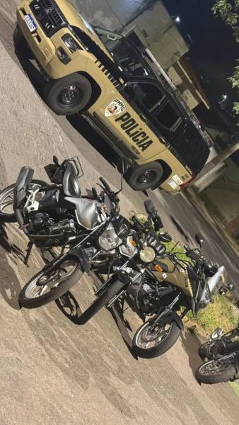 Uma viatura policial amarela estacionada ao fundo com a palavra 'POLÍCIA' visível, enquanto várias motos estão agrupadas em primeiro plano.