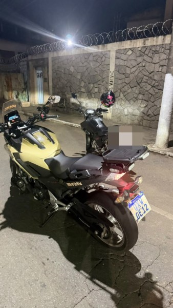 Motocicleta amarela na rua à noite, com uma segunda motocicleta ao fundo e um muro de pedras no cenário.
