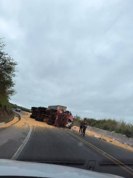 Caminhão tombado na estrada, com carga espalhada sobre a pista, e pessoas próximas inspecionando a situação.
