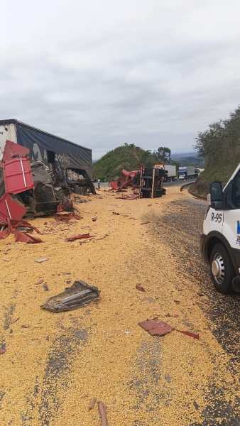 Caminhão tombado na estrada, com carga de milho espalhada pelo chão e destroços ao redor.
