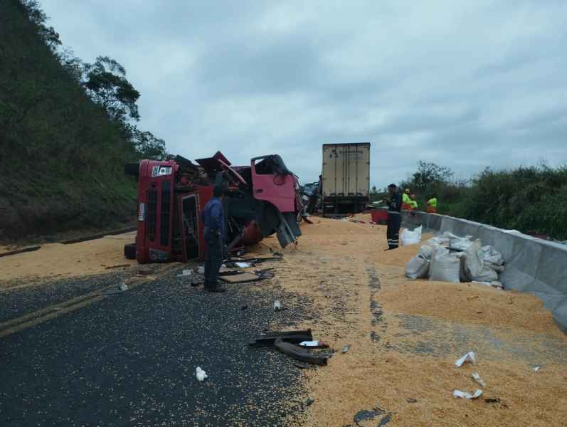 Caminhão tombado na estrada com grãos espalhados pela via, enquanto trabalhadores realizam limpeza.