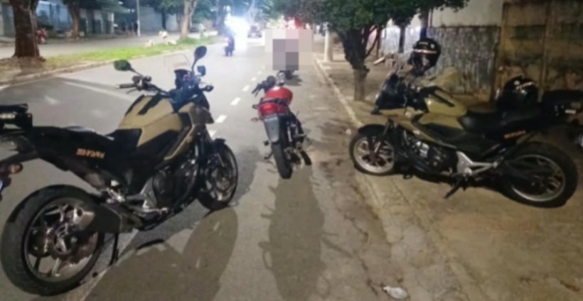 Duas motocicletas de cores escuras estacionadas na calçada à noite, com uma moto menor vermelha entre elas, em uma rua urbana.