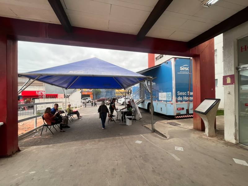 Área externa coberta com uma tenda azul, onde pessoas estão sentadas em cadeiras, conversando e sendo atendidas em uma van do Sesc.