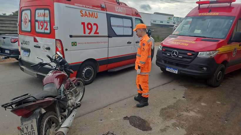 Um profissional de resgate em uniforme laranja observando uma motocicleta danificada em frente a ambulâncias do SAMU e dos bombeiros.