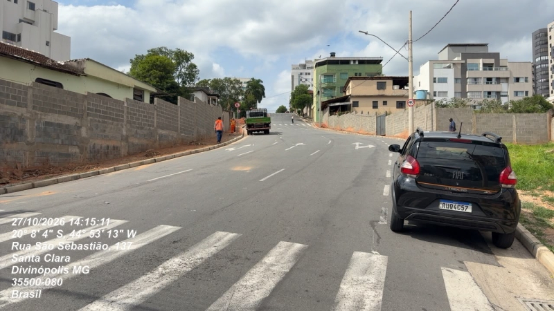 Vista de uma rua em Divinópolis, MG, com um carro estacionado à direita e trabalhadores em uma obra na calçada.