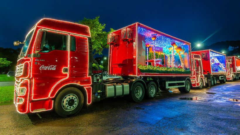 Caminhão vermelho da Coca-Cola decorado com luzes de Natal, estacionado à noite, com outros caminhões ao fundo.