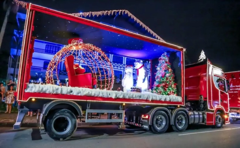 Caminhão decorado para o Natal, com luzes cintilantes, um cenário festivo contendo uma poltrona vermelha, ursos polares e uma árvore de Natal, em uma noite iluminada.