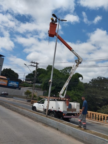 Trabalhador em um caminhão com guindaste, realizando manutenção em um poste de iluminação em uma rua, sob céu azul com nuvens.