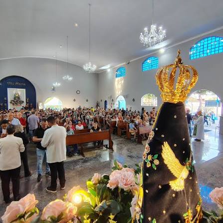 Vista interna de uma igreja cheia de pessoas durante um evento religioso, com um altar decorado e uma figura sagrada em destaque à frente.