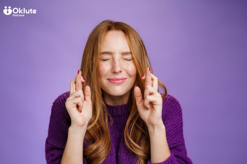 Mulher com cabelo ruivo, usando um suéter roxo, sorrindo e com os olhos fechados, fazendo um gesto de cruzar os dedos sobre um fundo roxo.